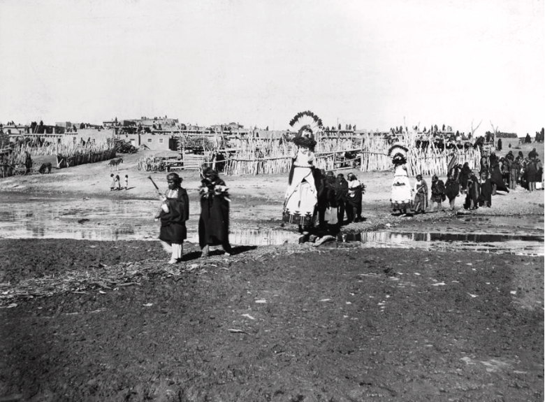 A black-and-white photograph of people dressed in ceremonial black clothes, marching in the mud and carrying tall white, feathered puppets.
