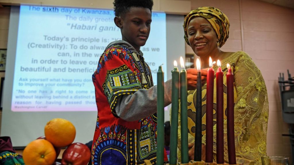 A black woman dressed all in yellow watches a black boy dressed in red light one of seven red, green and black candles. Behind them is a projector slideshow presentation.  