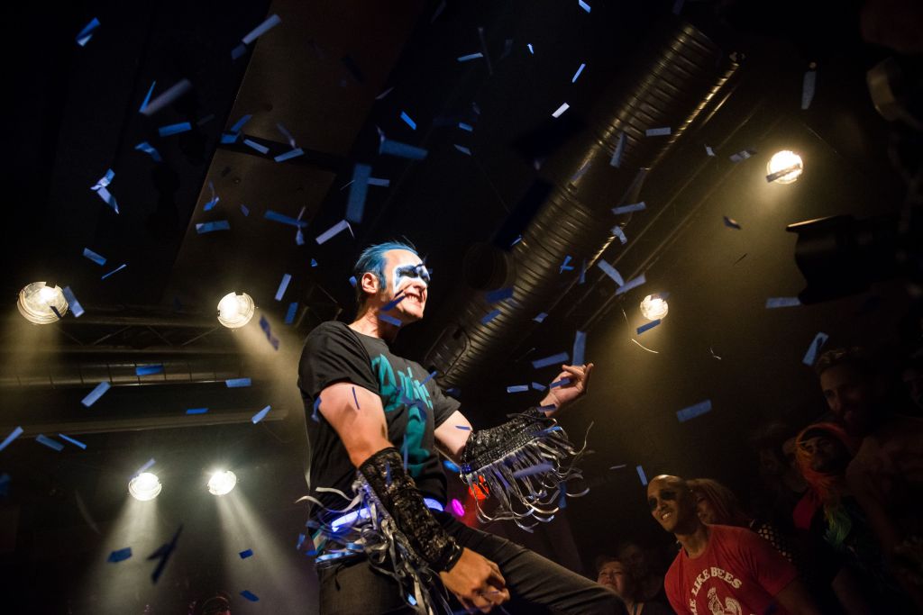 A man in a black shirt, pants, and frilled wrist gauntlets, with blue hair and eye makeup, surrounded by blue confetti, plays an invisible guitar.