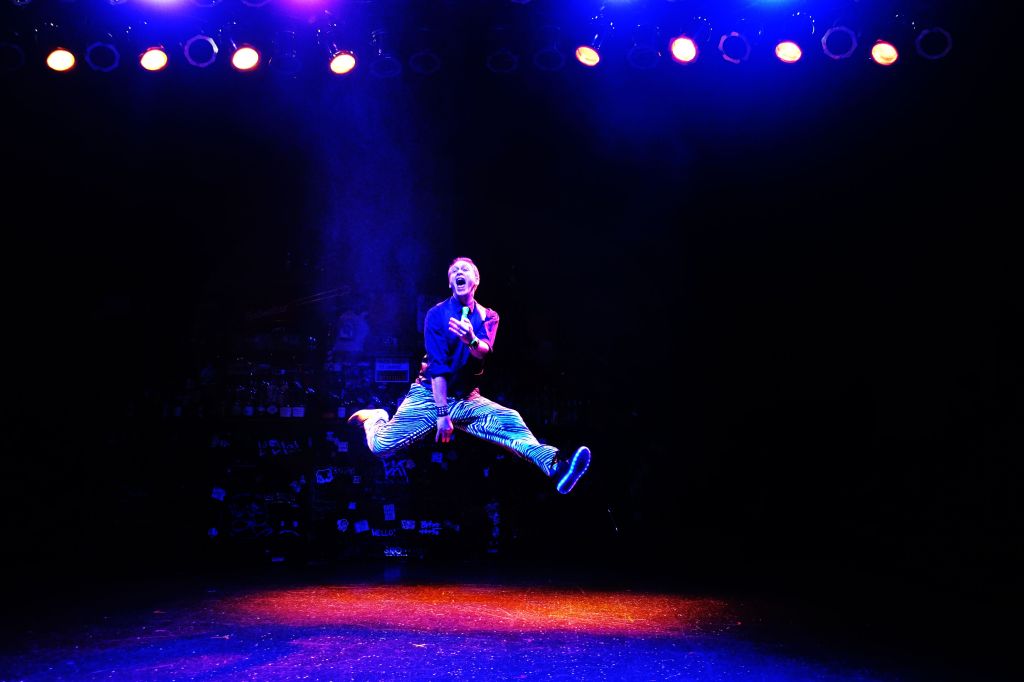 A man under blue and red lights jumping and doing a split, playing an invisible guitar.